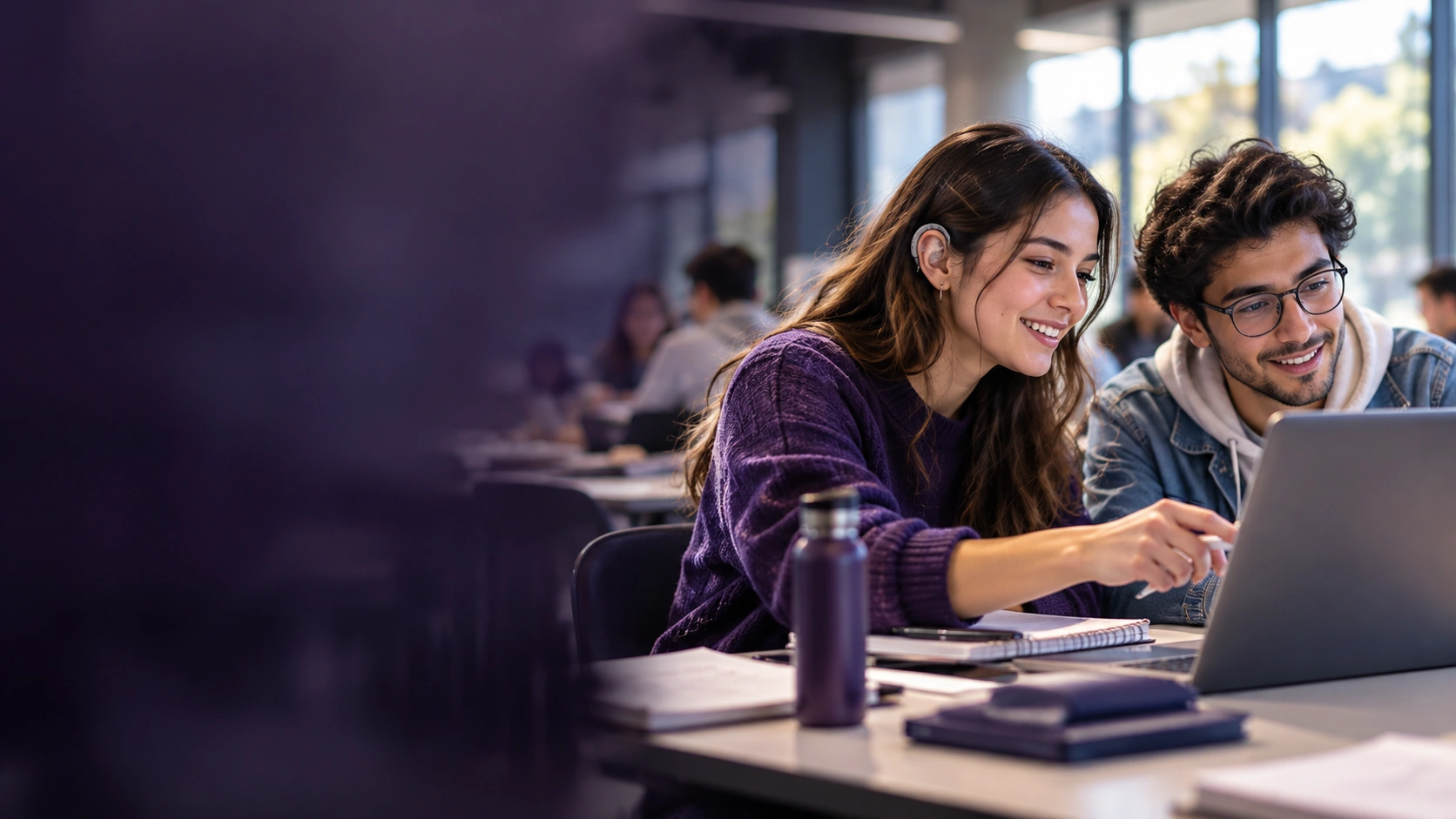 Dos estudiantes consultan un computador durante una clase, una de ellas utiliza un dispositivo auditivo